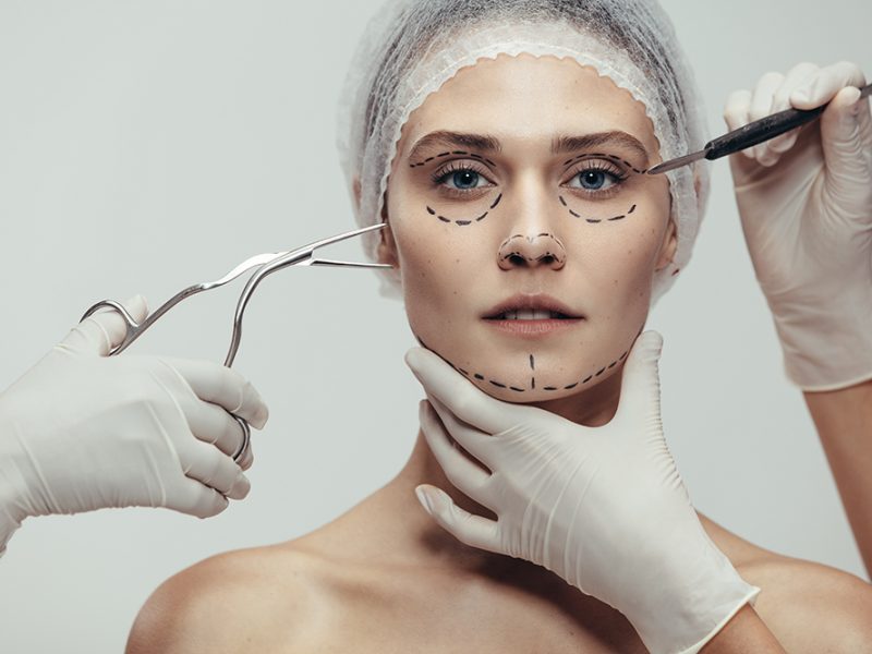 Close up of woman having cosmetic face surgery with scalpel and medical forceps against grey background. Woman having a anti aging and face lift surgery.